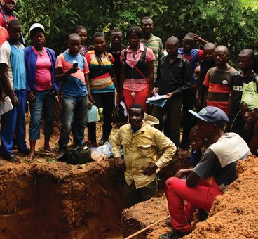 A photo of Esakakondo "Al" Lohese standing in a soil pit with several people listening around him.